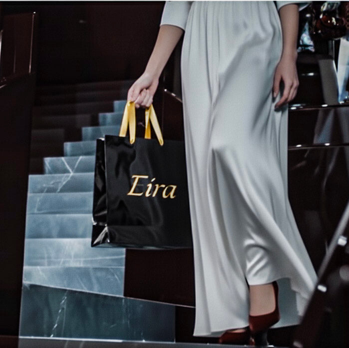 A woman wearing a long white dress while holding a black shopping bag with 'Eira' branding while stepping down on marble steps.